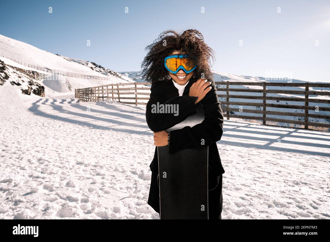 Happy Ethnic Sportswoman dans une tenue élégante et des lunettes de protection embrassant le snowboard sur un support enneigé en plein soleil Banque D'Images