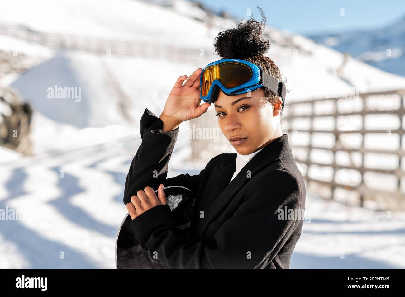 Jolie athlète afro-américaine en tenue tendance et protectrice lunettes debout avec snowboard regardant la caméra en hiver Banque D'Images