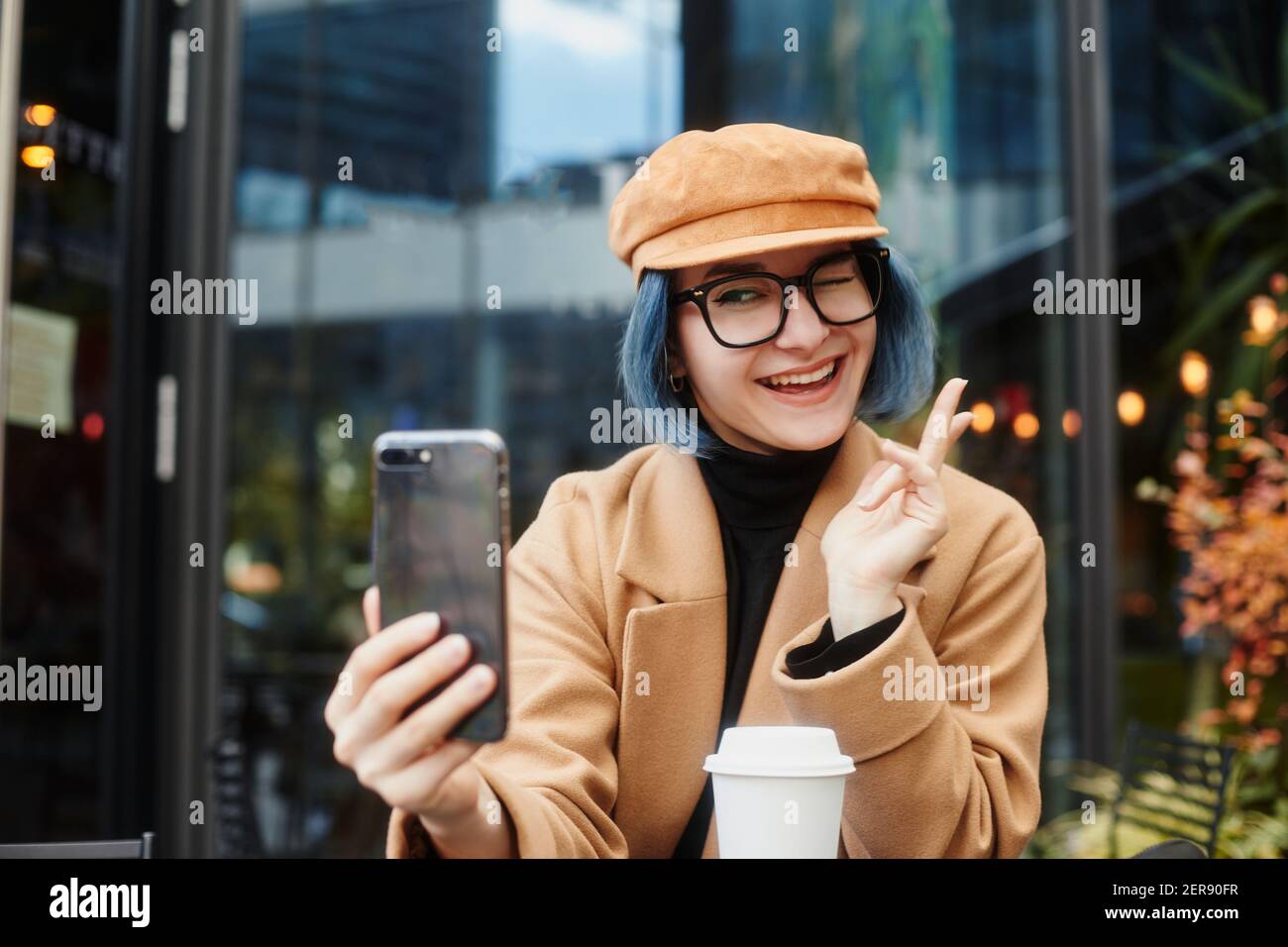 Une fille avec des cheveux bleus et des lunettes est assise à une table dans un café dans la rue. Tenir une tasse de café et prendre des selfies. Banque D'Images