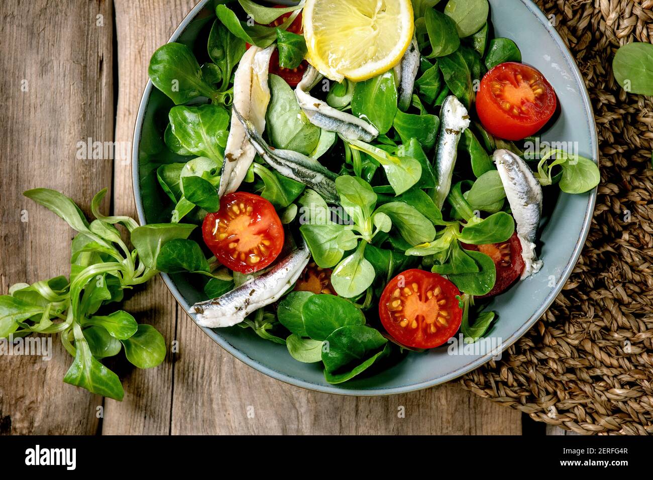 Salade verte avec filet d'anchois ou de sardines marinés et tomates  cerises, servies dans un bol bleu avec du citron et de l'huile d'olive sur  une serviette en paille au-dessus de l'ol