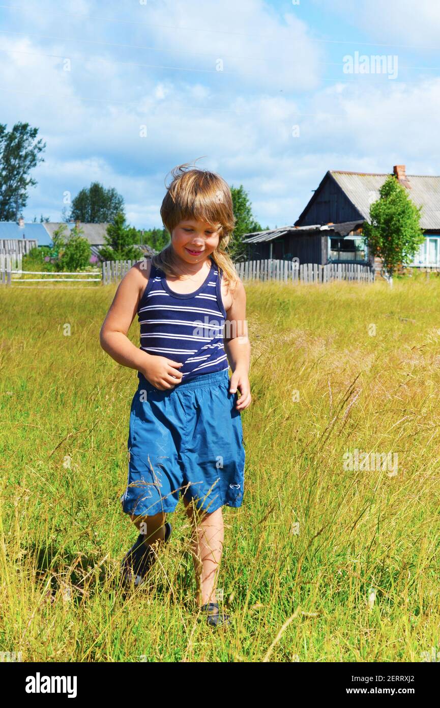 Un garçon en short bleu et un t-shirt rayé se promène dans la prairie. Maisons de village à l'arrière. L'enfant sourit. Bonne humeur. Une enfance insouciante. Banque D'Images