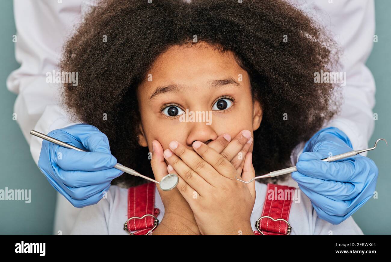 Petite fille afro-américaine effrayée à la rendez-vous du dentiste et couvre sa bouche avec les mains tout en examinant les dents Banque D'Images
