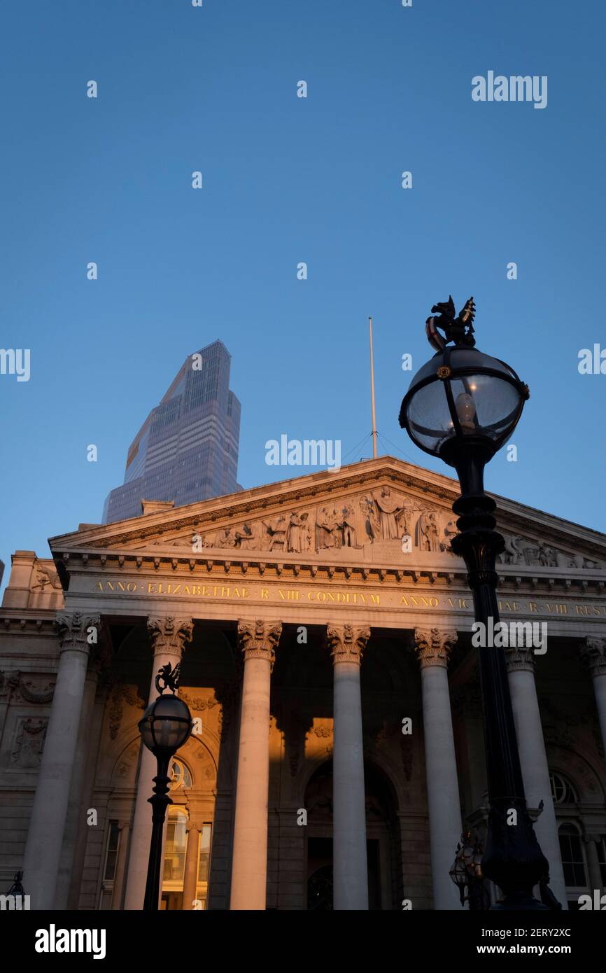 Avec des bureaux modernes d'institutions financières derrière, une vue panoramique architecturale sur les frises et inscriptions latines sur le fronton de la Bourse royale dans la ville de Londres, le quartier financier de la capitale, le 27 février 2021, à Londres, en Angleterre. Au sommet des colonnes doriques et ioniques avec leur travail en pierre, des linteaux puissants se croisent, portant la charge de l'art et des sculptures fines qui présentent le design de Sir William Tite en 1842-1844 et ouvert en 1844 par la reine Victoria dont le nom est écrit en latin (victoriae R). C’est le troisième bâtiment du genre érigé sur la sa Banque D'Images