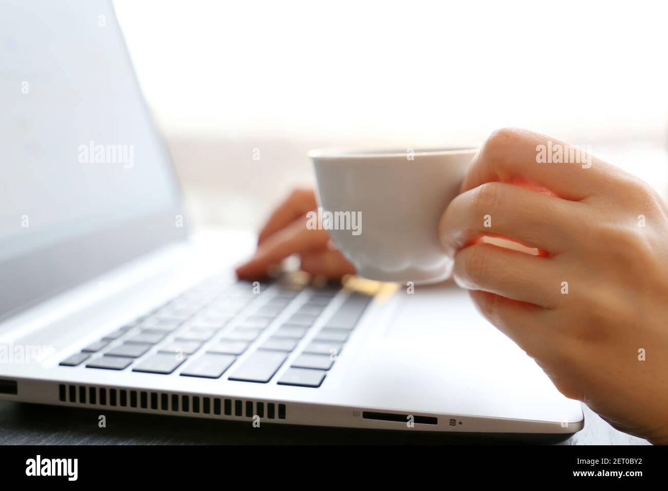 Une femme boit du café assis au clavier de l'ordinateur portable sur fond de fenêtre. Tasse blanche de boisson chaude dans la main de la femme au soleil, Bonjour Banque D'Images
