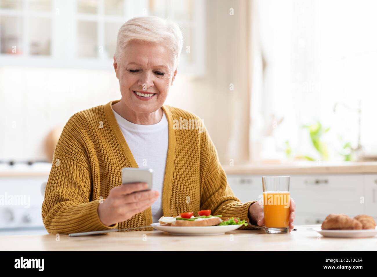 Femme âgée souriante utilisant un téléphone portable tout en déjeunant Banque D'Images