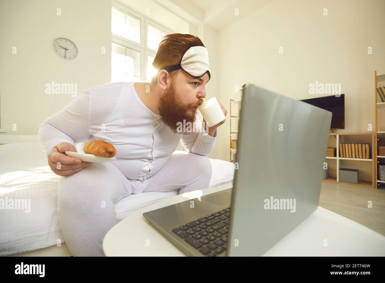 Drôle de barbu surpoids homme assis avec café et un petit pain pour petit déjeuner et regarder des films sur un ordinateur portable Banque D'Images