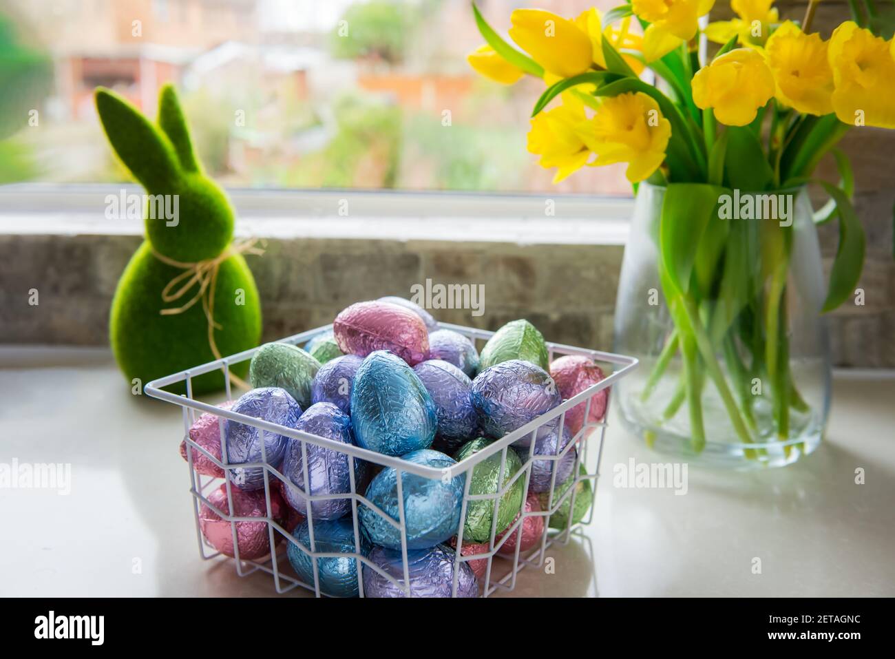 Panier en métal avec œufs de pâques au chocolat en feuilles de couleur, bouquet de tulipes jaunes et fleurs de jonquilles et lapin vert de Pâques sur table de cuisine blanche Banque D'Images