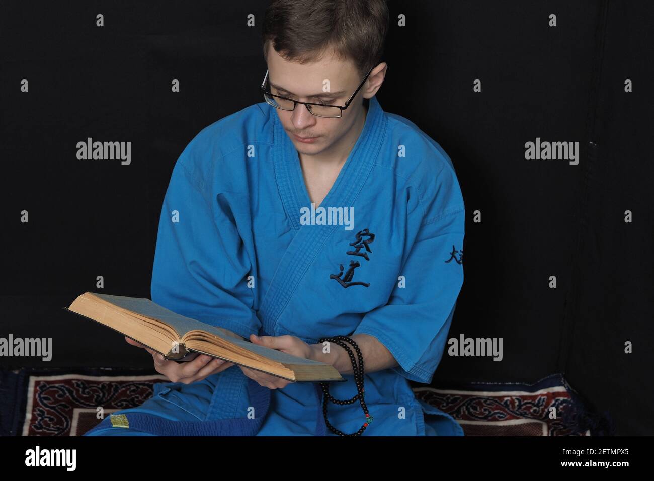 Un jeune homme avec des lunettes lit un livre assis sur le sol. Banque D'Images