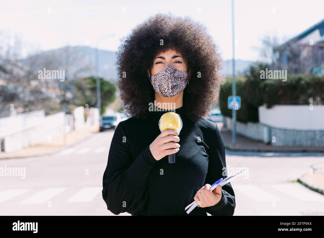 Journaliste de télévision féminine aux cheveux d'afro, portant un masque de peau léopard pour se protéger d'un virus, racontant une histoire dans la rue Banque D'Images