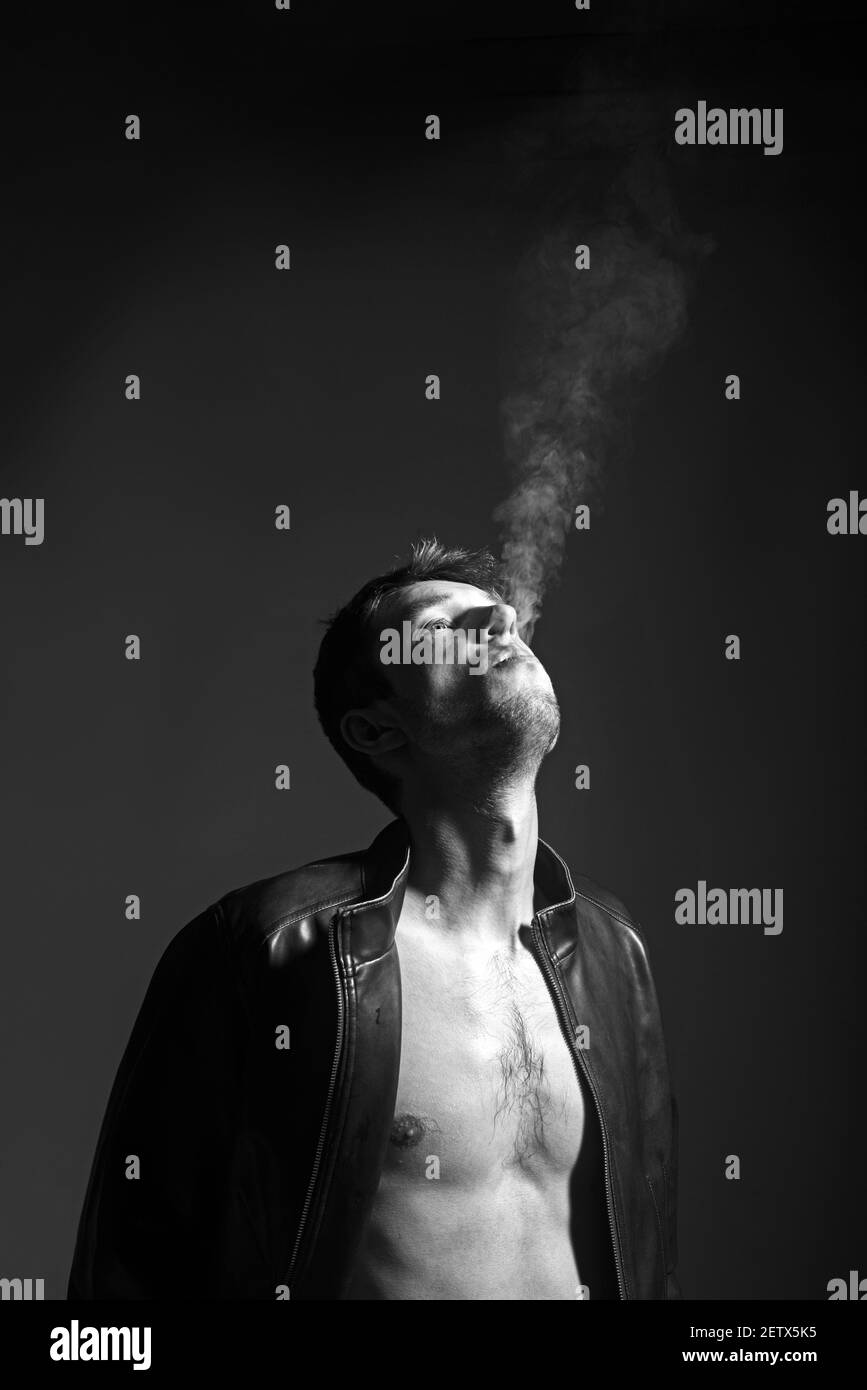 Portrait d'un jeune homme brutal et sexy dans une veste en cuir et nu torse exhales fumée vers le haut. Banque D'Images