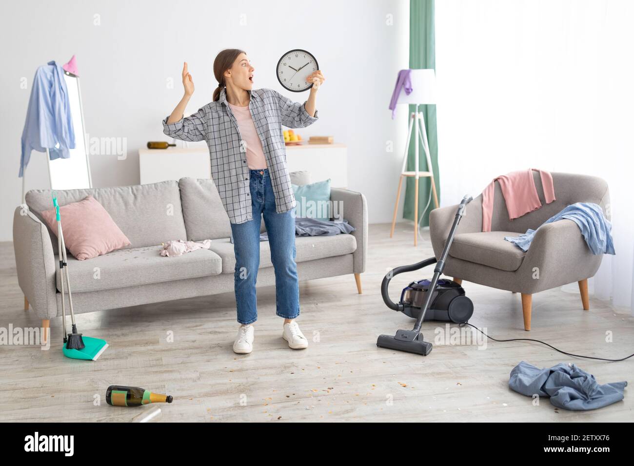 Une jeune femme choquée tient une horloge, se sentant terrifiée, debout dans une pièce désordonnée après la fête, étant en retard pour nettoyer l'appartement Banque D'Images