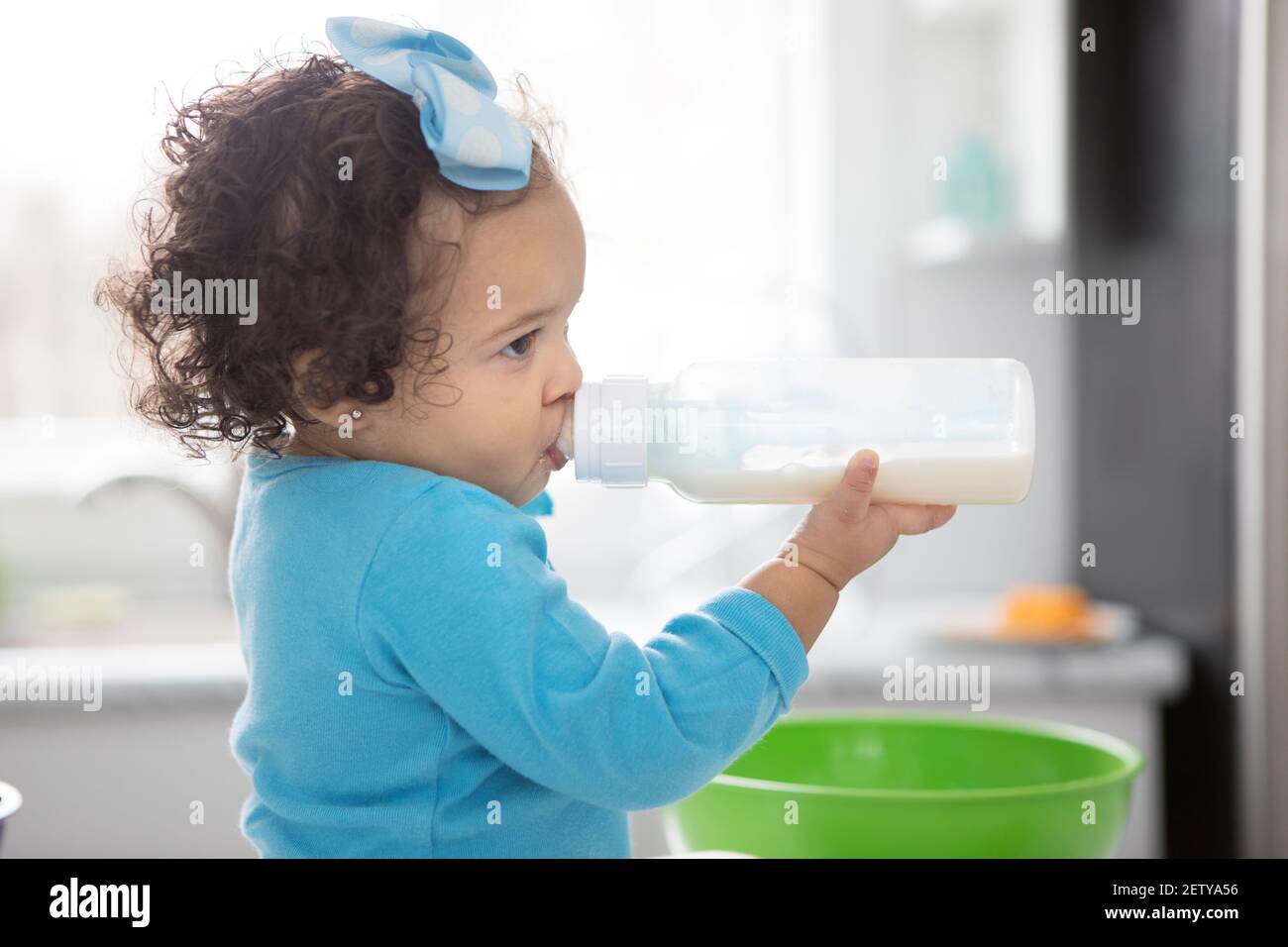 Une petite fille boit du lait dans une bouteille en plastique dans une cuisine moderne et lumineuse. Banque D'Images