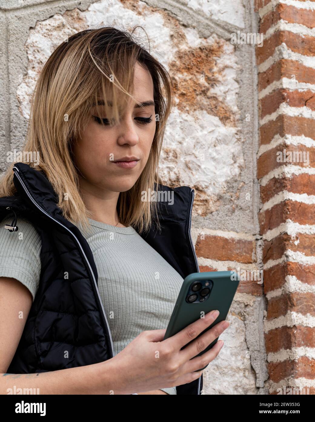 Jolie jeune fille blonde consulte les notifications sur son mobile avec trois caméras et un étui vert, pendant qu'elle attend à côté d'un mur de pierre Banque D'Images