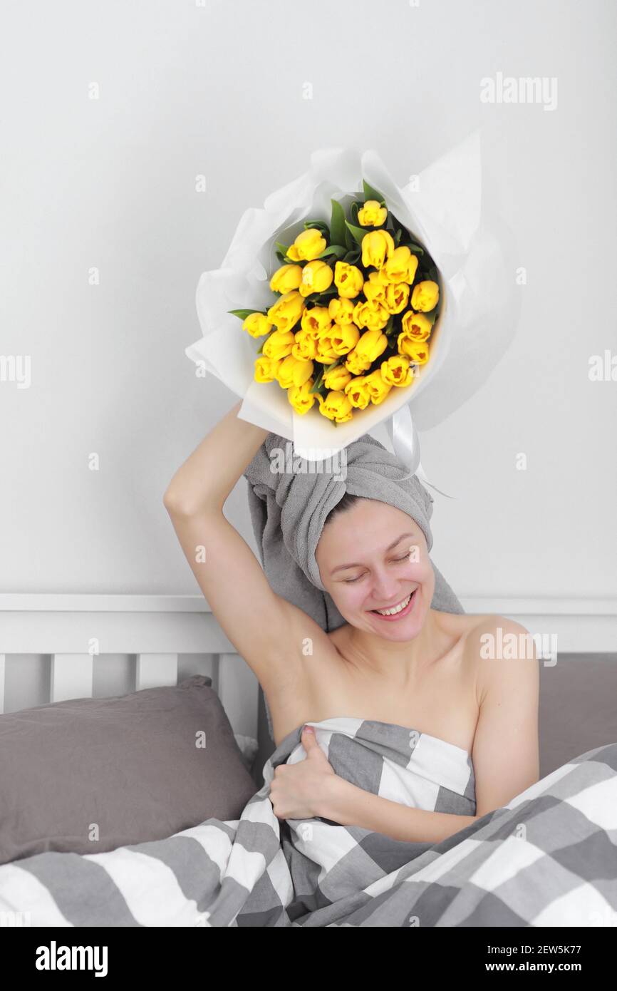 femme heureuse avec des fleurs de tulipe jaunes dans le lit après la douche avec une serviette sur la tête appréciant cadeau romantique pour la saint valentin. lui a donné un bouquet de fleurs. Banque D'Images