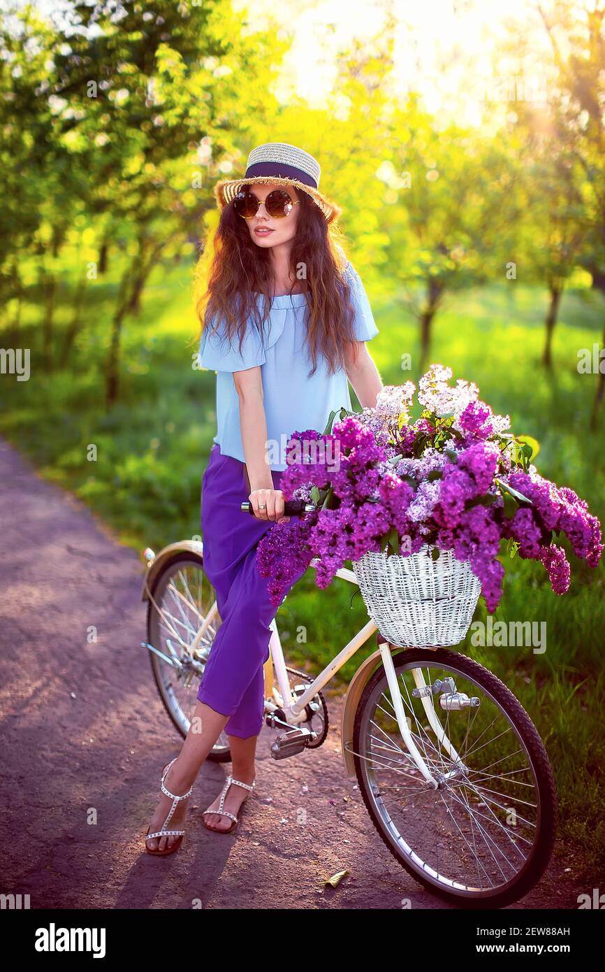 Portrait d'une jeune fille heureuse avec vélo vintage et fleurs sur fond de ville en plein soleil. Vélo avec panier rempli de fleurs. Concept de loisirs actifs. Banque D'Images