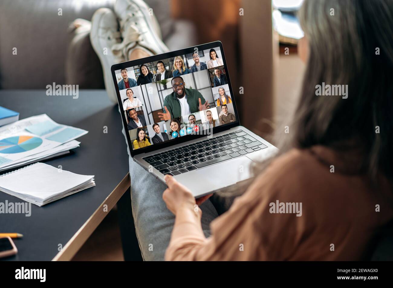 Briefing en ligne. Une femme moderne et élégante, aux cheveux gris mûrs, communique avec ses collègues en ligne via un appel vidéo, utilise une application et un ordinateur portable, assise à son bureau à domicile et jetant ses pieds sur la table Banque D'Images