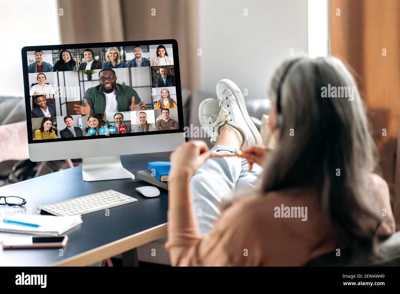 Une femme moderne mûre grise communique avec ses collègues en ligne par vidéoconférence utilise l'ordinateur, assis à son bureau à domicile, jetant ses pieds sur la table, sur l'écran de nombreuses personnes différentes Banque D'Images