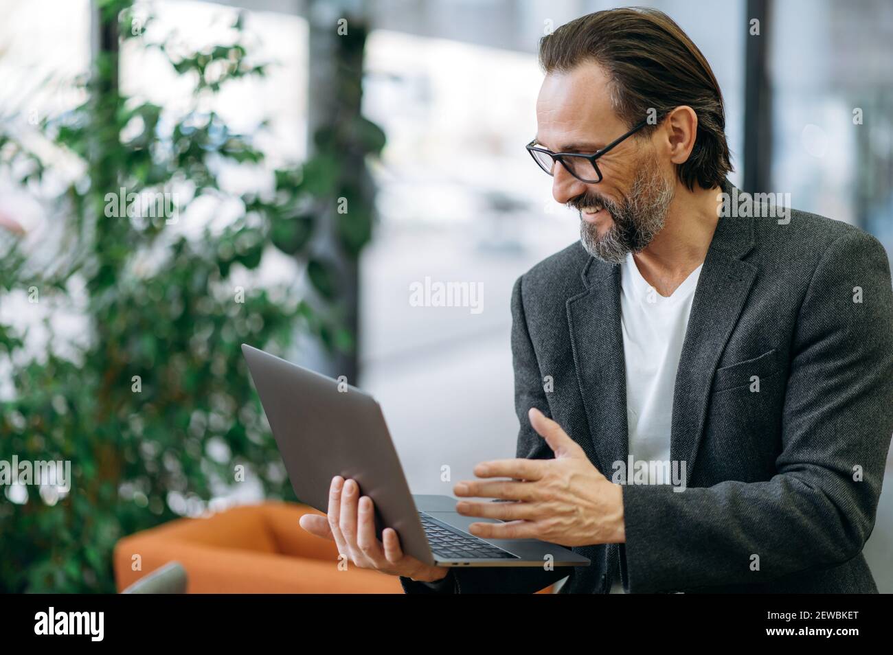 Homme d'affaires plein de joie lors de visioconférences avec des collègues, discutant d'un projet d'entreprise. Beau employé de sexe masculin utilisant un ordinateur portable, assis sur un canapé dans un bureau moderne, souriant Banque D'Images