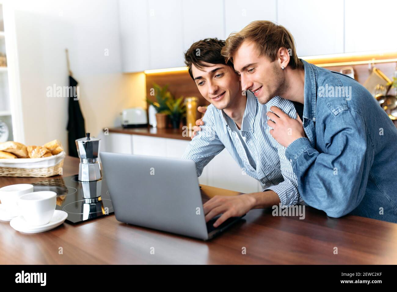 Deux homosexuels heureux. Gay couple debout dans la cuisine, utiliser un ordinateur portable, naviguer sur Internet, regarder un film, faire du shopping en ligne, passez du temps ensemble Banque D'Images