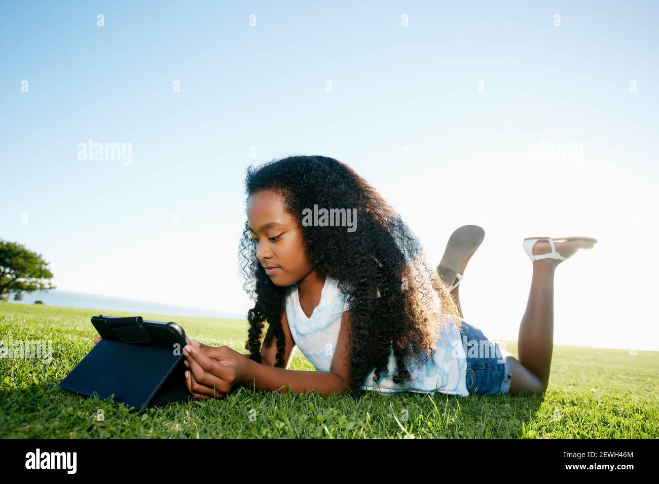Jeune fille de neuf ans de race mixte couché sur l'herbe regardant une tablette numérique. Banque D'Images