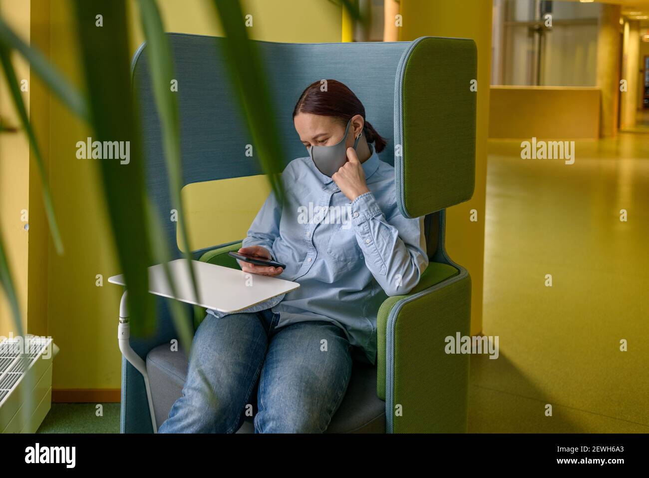 Femme en masque de détente sur chaise confortable dans le coin du bureau ou de l'université avec téléphone. Distanciation sociale, isolement, solitude, seul Banque D'Images