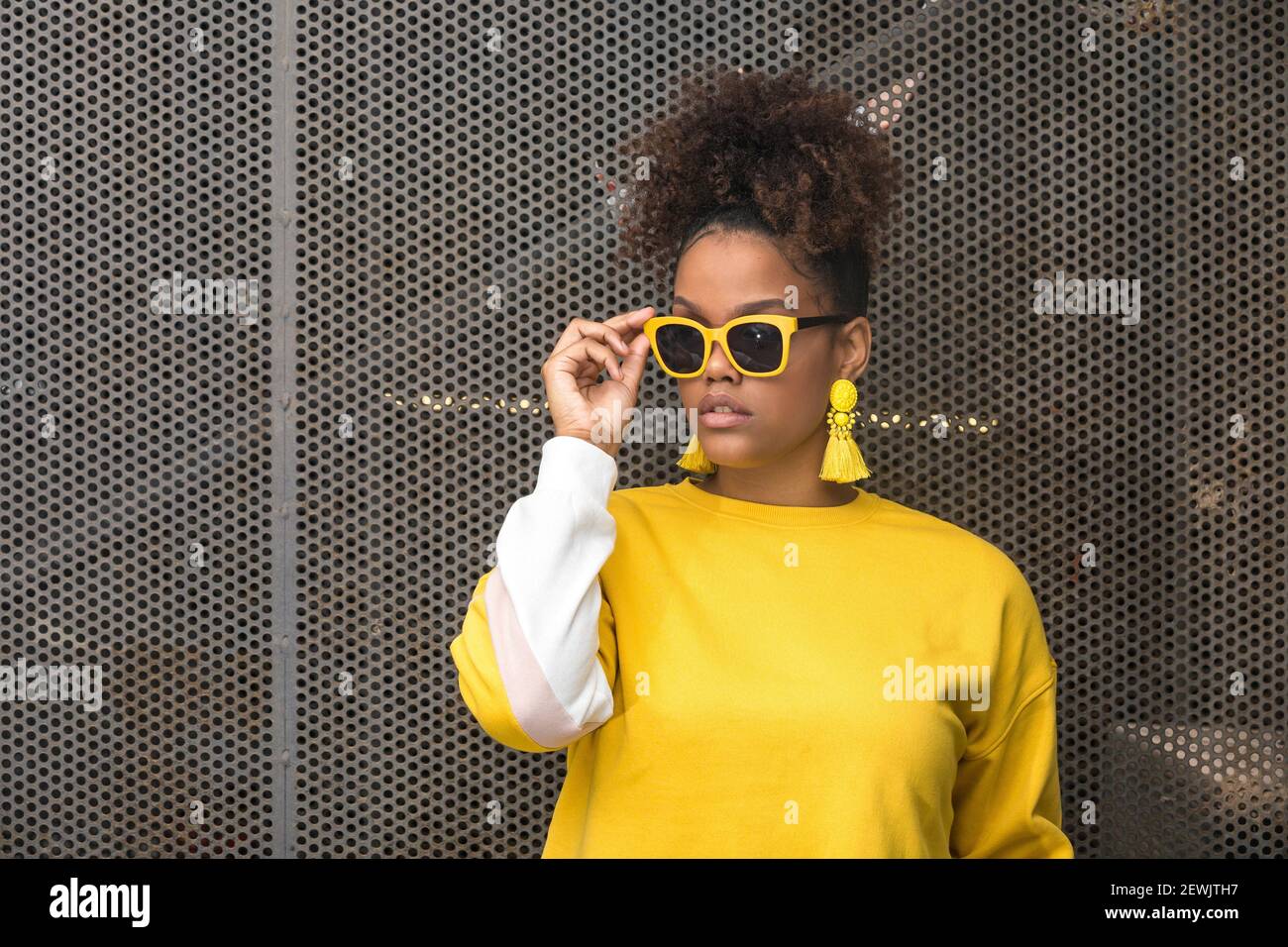 Jeune femme afro-américaine à la mode et confiante, ornée d'un chemisier  jaune vif et boucles d'oreilles qui ajustent les lunettes de soleil tendance  tout en se tenant contre le mesh Photo Stock -