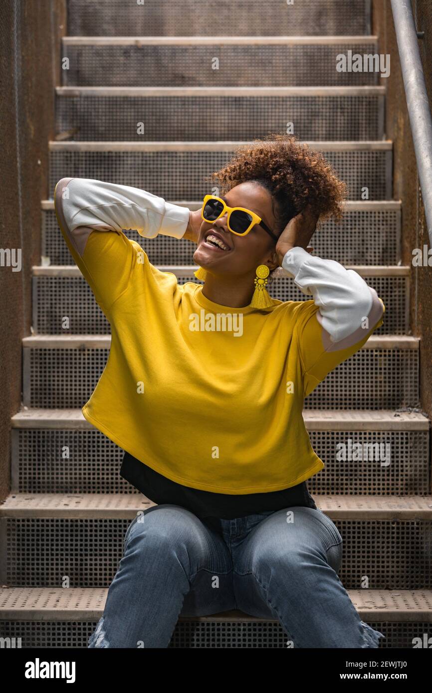 Jeune femme afro-américaine joyeuse en chemise jaune vif tendance et des  lunettes de soleil avec boucles d'oreilles ayant plaisir et touchant la  tête pendant assis Photo Stock - Alamy