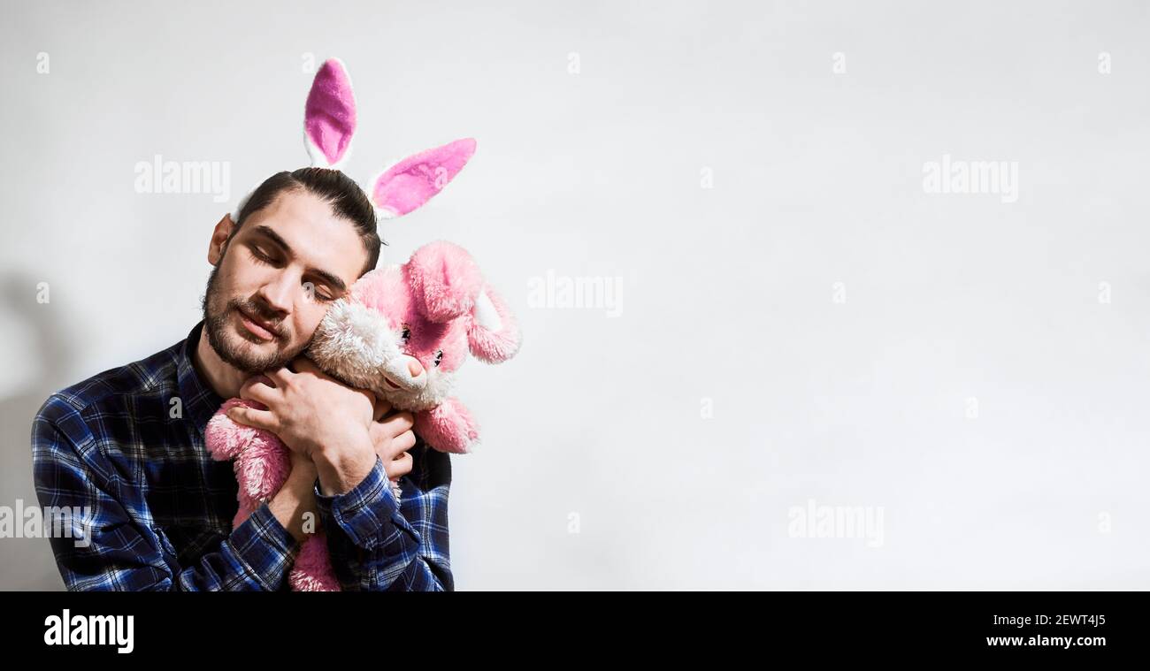 Charmant jeune homme caucasien avec la barbe et le lapin rose des oreilles de Pâques câline le lapin farci serré et ferme ses yeux dans le plaisir. Bannière avec emp Banque D'Images