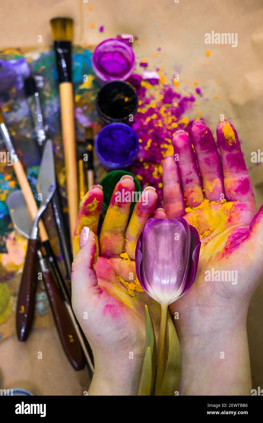 Fleur de tulipe dans la main aquarelle du peintre. Les outils de coloration sont sur la table, les bocaux avec des couleurs et de la poudre sont dans l'atelier. Banque D'Images
