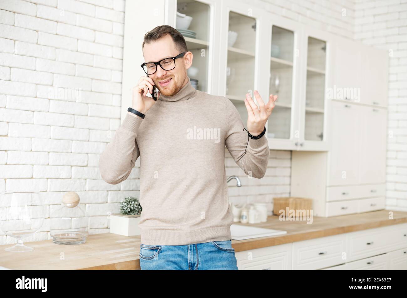 Charmant homme d'affaires hippster barbu dans des vêtements confortables et décontractés dans des lunettes debout dans la cuisine tenant un téléphone mobile et parlant avec une petite amie, famille, client, traitant des problèmes de client Banque D'Images