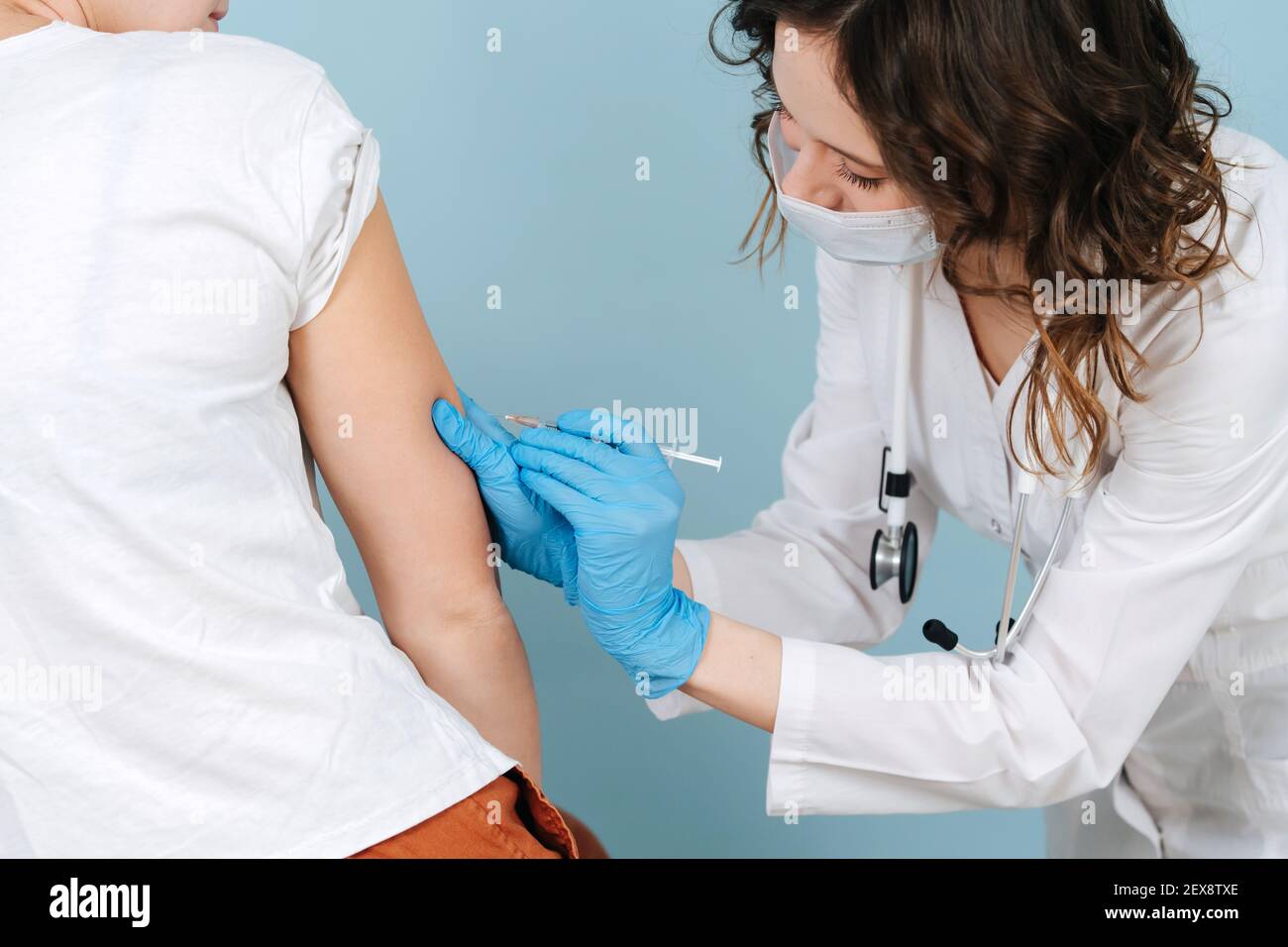 Infirmière spécialisée qui donne l'injection du vaccin à l'épaule. Sur fond bleu. Elle porte une robe blanche et un masque médical. Banque D'Images