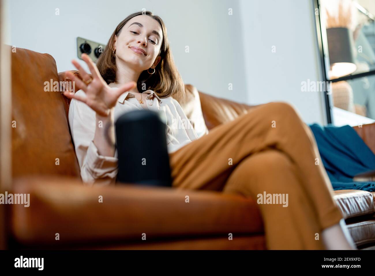 Jolie femme heureuse parlant avec la colonne d'assistant audio noire sur la table basse tout en étant assise sur un canapé en cuir marron dans le salon élégant. Concept de maison intelligente. Banque D'Images