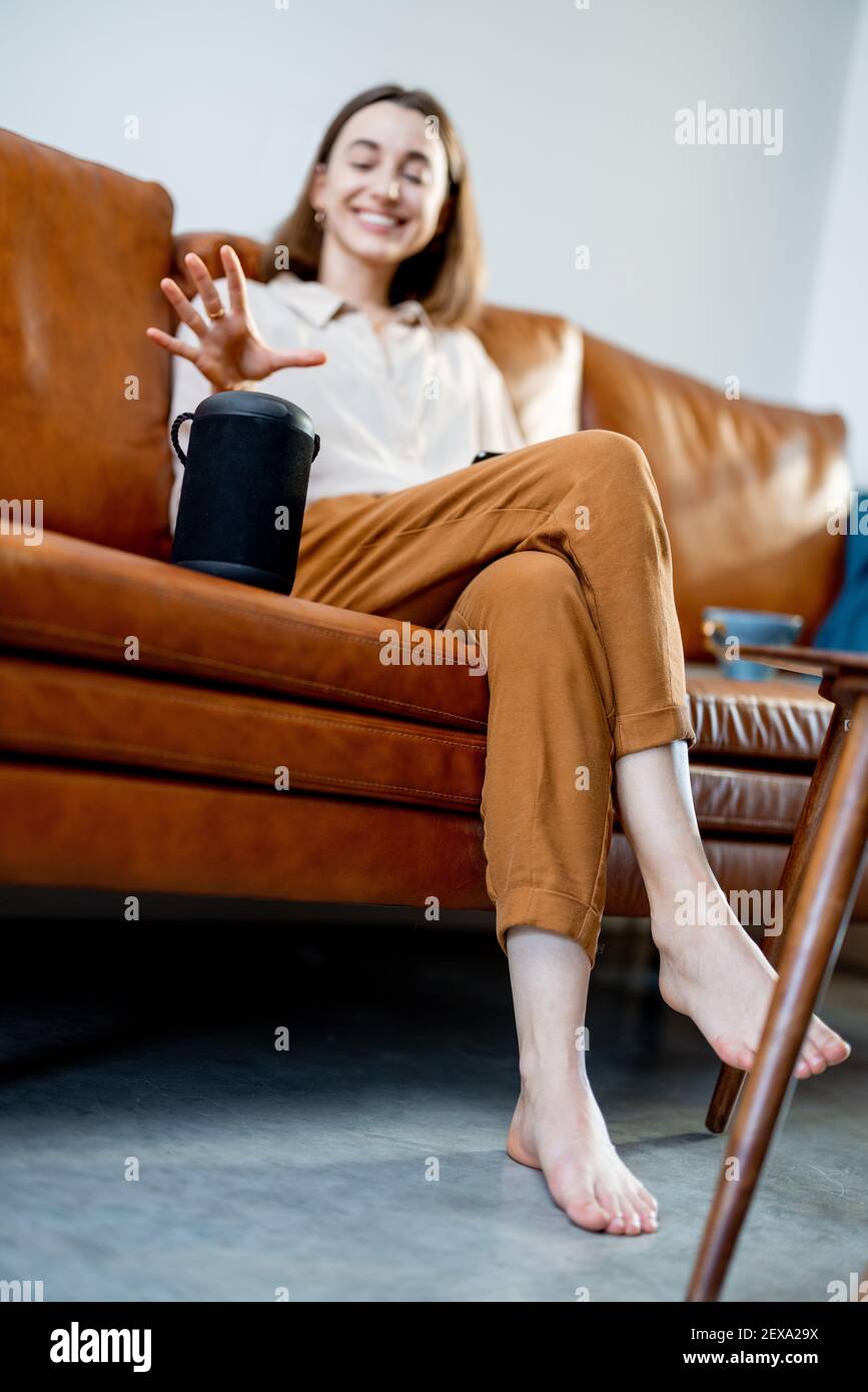 Jolie femme heureuse parlant avec la colonne d'assistant audio noire sur la table basse tout en étant assise sur un canapé en cuir marron dans le salon élégant. Concept de maison intelligente. Banque D'Images