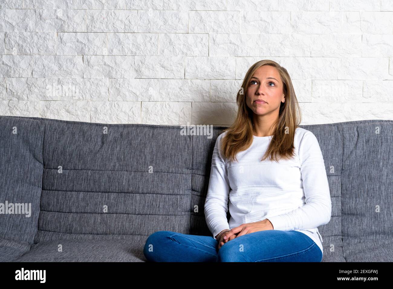 Femme caucasienne assis soigneusement sur le canapé devant le blanc mur de pierre et regarder vers le haut Banque D'Images