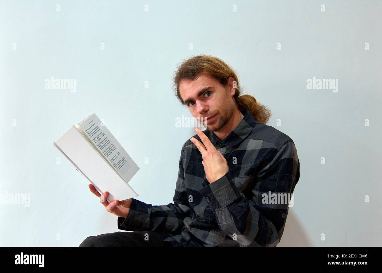 Un homme caucasien tenant un livre et montrant la paix sur l'arrière-plan d'un mur blanc Banque D'Images