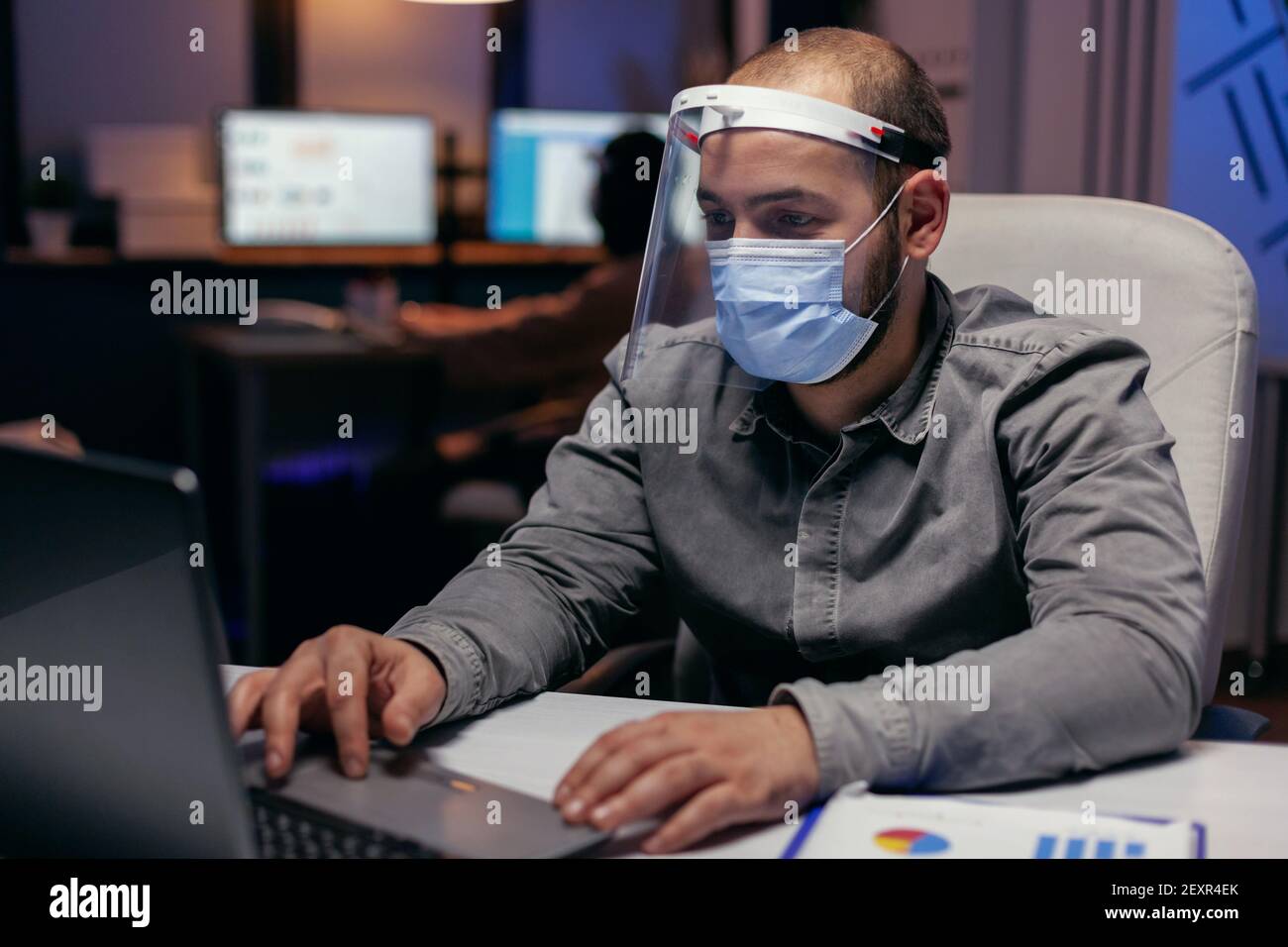 L'homme d'affaires se protège avec la visière contre le covid-19 tard dans le bureau distancer de ses collègues. Défilement sur ordinateur portable. Homme stressé en société travaillant dur pour terminer un projet portant un masque facial comme précaution de sécurité en raison d'une pandémie de coronavirus. Banque D'Images