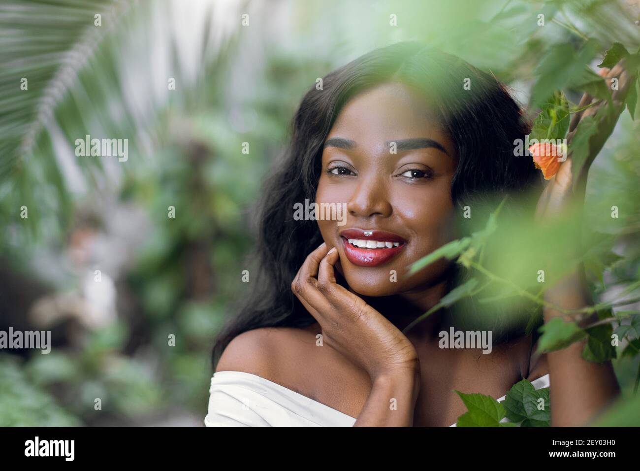 Portrait d'une jeune fille africaine charmante et exotique, avec des cheveux longs et foncés, regardant l'appareil photo tout en se tenant au milieu des feuilles de palmier et d'arbre à fleurs Banque D'Images