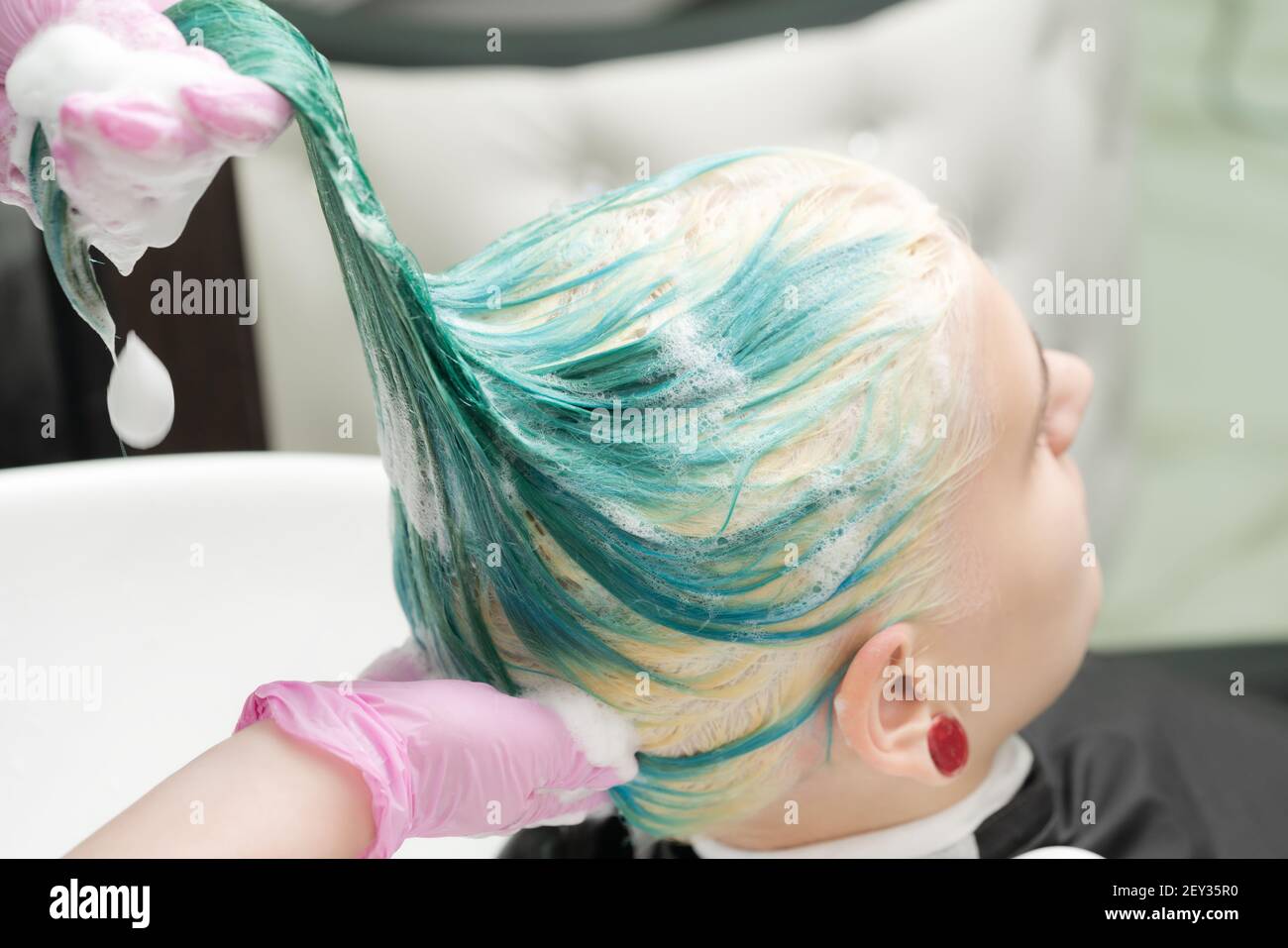 Laver les cheveux verts couleur de la jeune femme avec shampooing dans l'évier. Coiffeur de travail avec gants de protection roses dans un salon de beauté. Banque D'Images