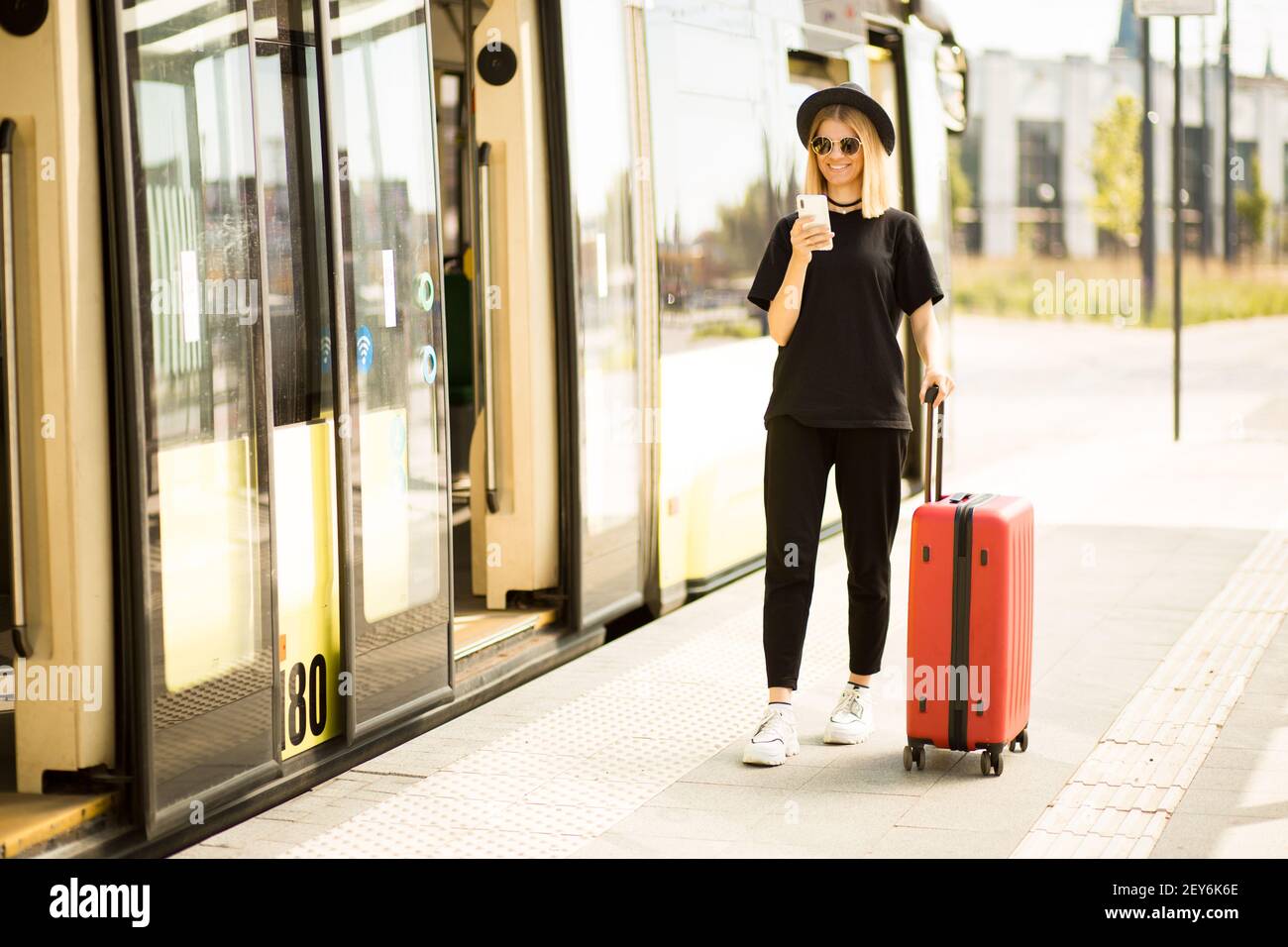 Voyageur touriste femme en noir vêtements décontractés, lunettes de soleil et chapeau avec valise rouge utiliser le téléphone mobile pour acheter des billets en ligne et s'asseoir à la gare routière. Les femmes voyagent à l'étranger le week-end pour une escapade. Concept de vol Banque D'Images