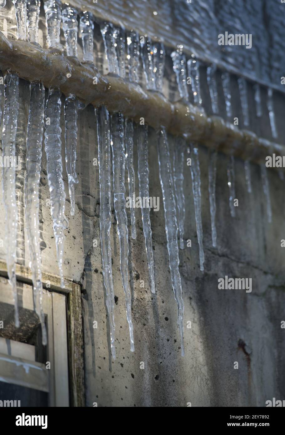 des glaçons pointus congelés suspendus à des tuyaux en métal industriel sur de l'eau grungie ont endommagé les ombres à glace du bâtiment en béton sur le mur extérieur en béton en arrière-plan Banque D'Images