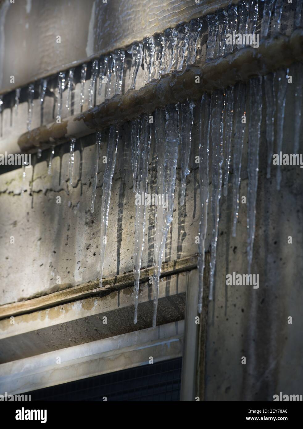 des glaçons pointus congelés suspendus à des tuyaux en métal industriel sur de l'eau grungie ont endommagé les ombres à glace du bâtiment en béton sur le mur extérieur en béton en arrière-plan Banque D'Images
