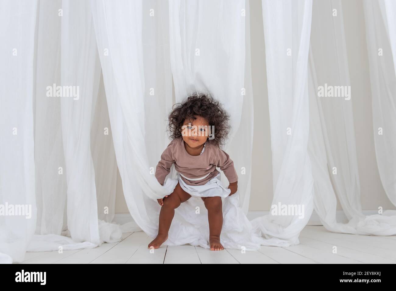 Petite fille américaine africaine bourieuse danse rires fun parmi le tissu de tulles blanc aéré. Intérieur minimaliste, style scandinave. Bonne enfance. Le Banque D'Images