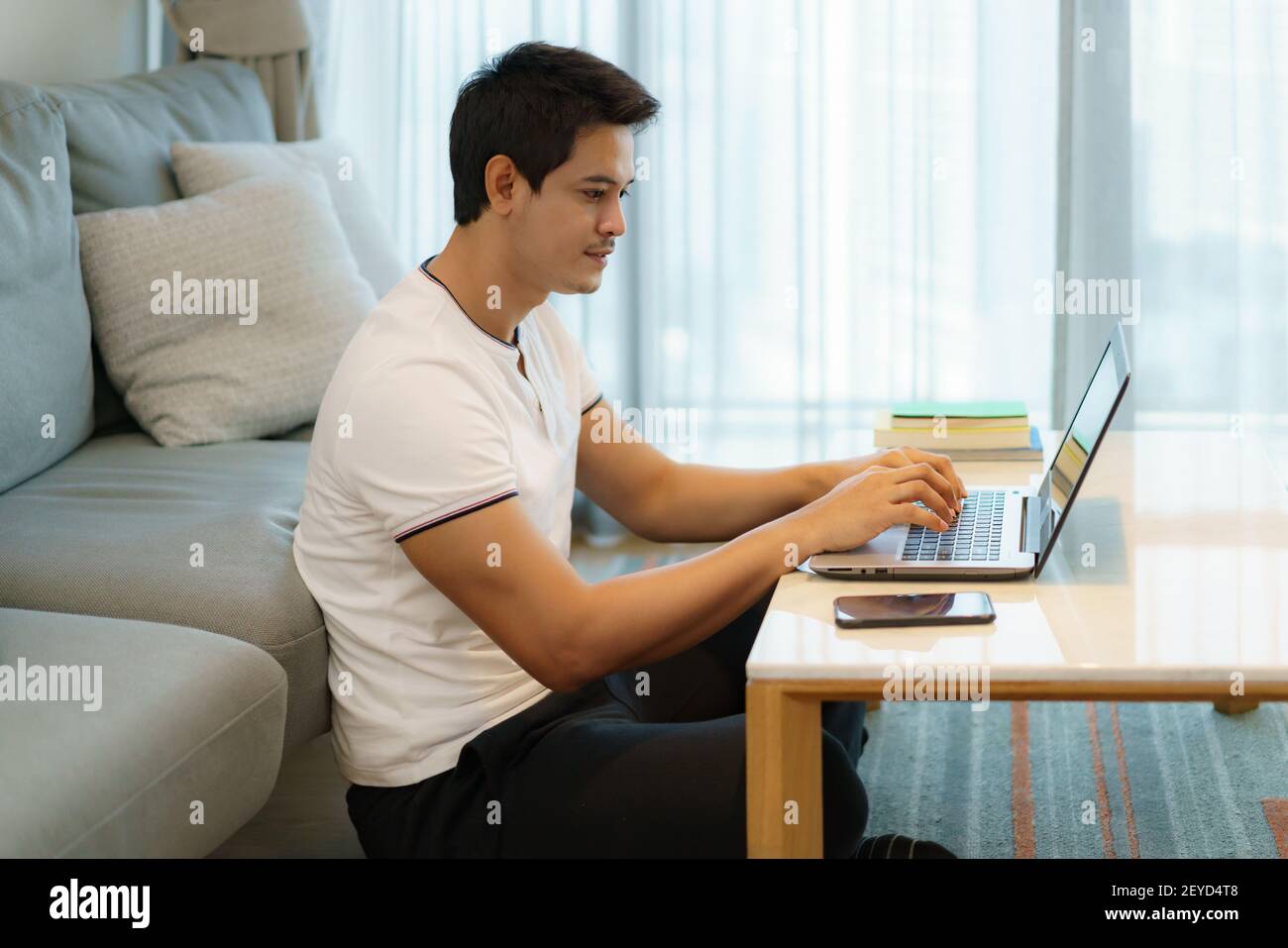 Un homme asiatique travaillant à la maison, discutant en ligne avec un collègue sur son ordinateur portable tout en étant sur le canapé dans le salon à la maison. Banque D'Images