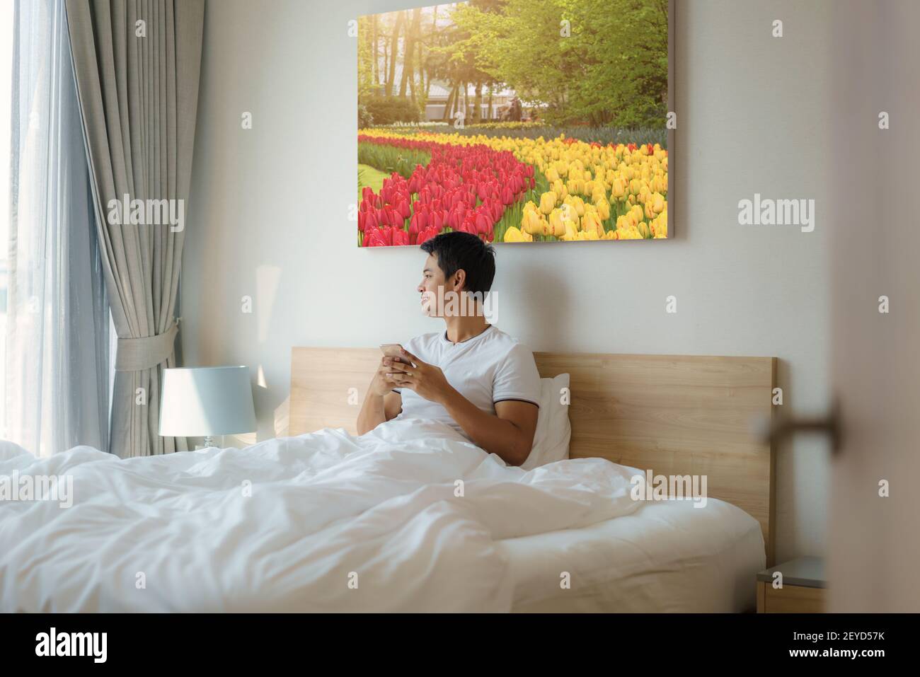 Un homme asiatique s'assoit au lit avec une couverture blanche et vérifie son e-mail sur son téléphone portable le matin à la maison avant de prendre une douche. Banque D'Images
