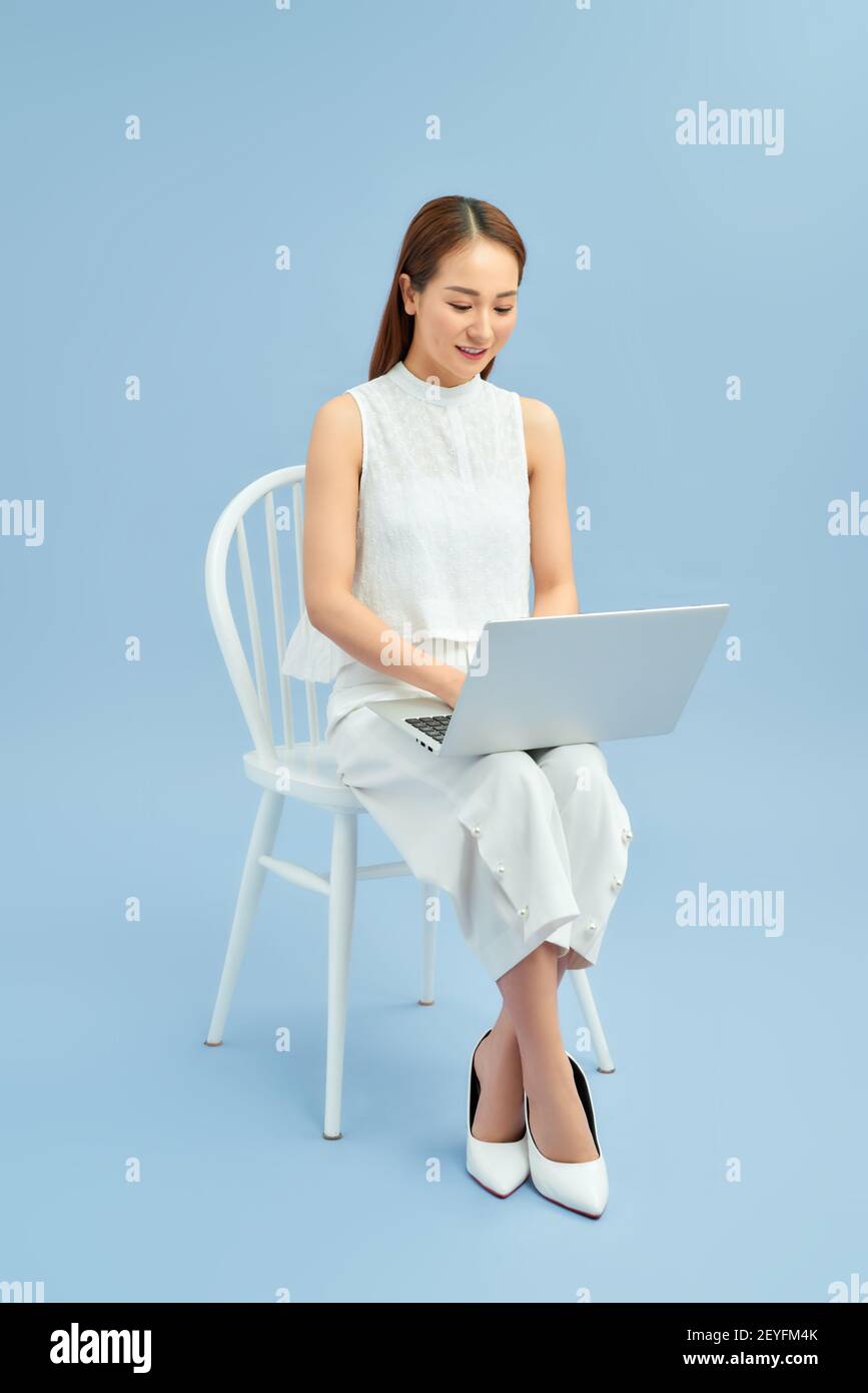 Jeune fille à la mode assise sur une chaise avec ordinateur portable isolé sur fond bleu. Oups ! Banque D'Images