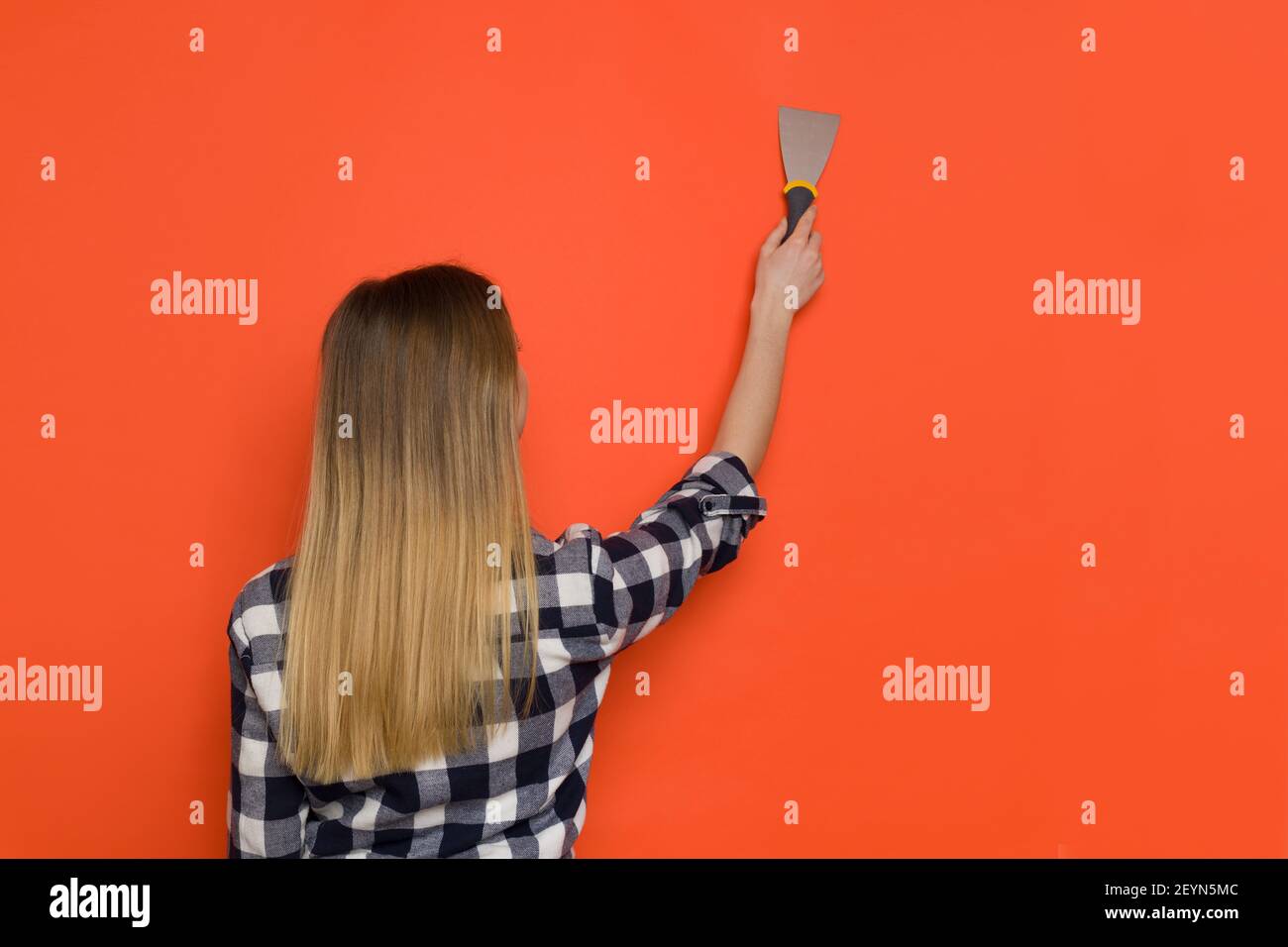 Jeune femme Blonde en maillot Lumberjack travaille avec la spatule de peintre sur le mur orange. Vue arrière. Prise de vue à la taille. Banque D'Images