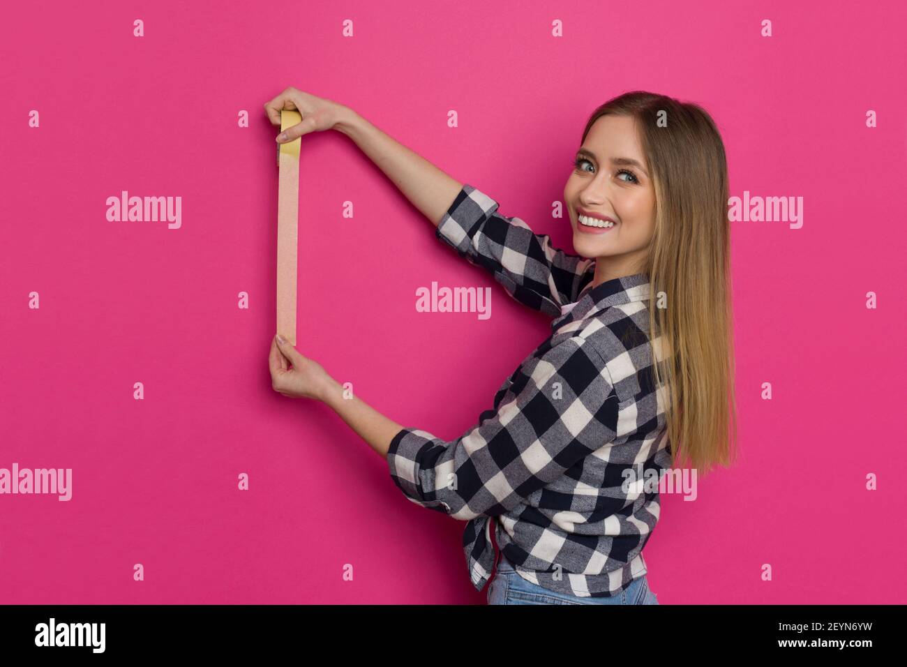 Une jeune femme blonde souriante en chemise de bûcheron tient la bande de peintre jaune sur le mur rose et regarde la caméra. Taille haute. Banque D'Images