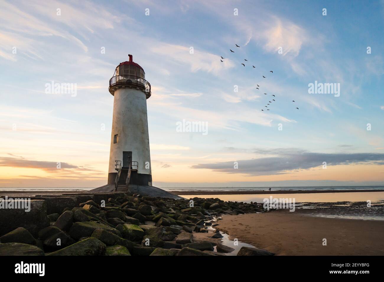 Magnifique phare au bord de la mer Golden hour coucher de soleil avec des rochers menant à des escaliers comme la marée de l'océan arrive. Oiseaux qui volent vers l'horizon, le soleil réfléchit l'eau Banque D'Images