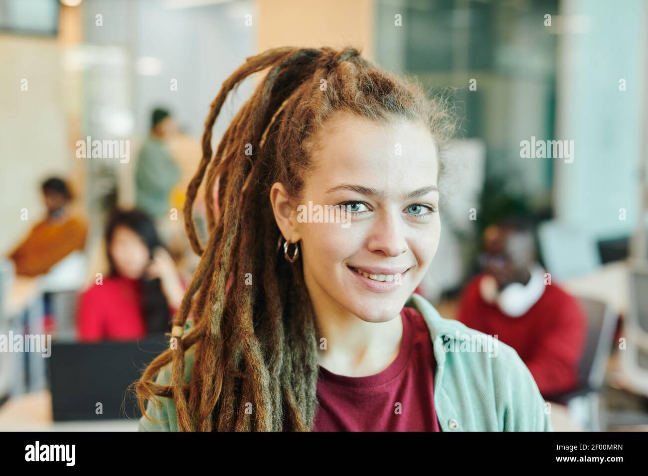 Jeune femme d'affaires souriante ou designer de vêtements décontractés debout devant de caméra contre ses collègues interculturels travaillant dans l'espace ouvert Banque D'Images