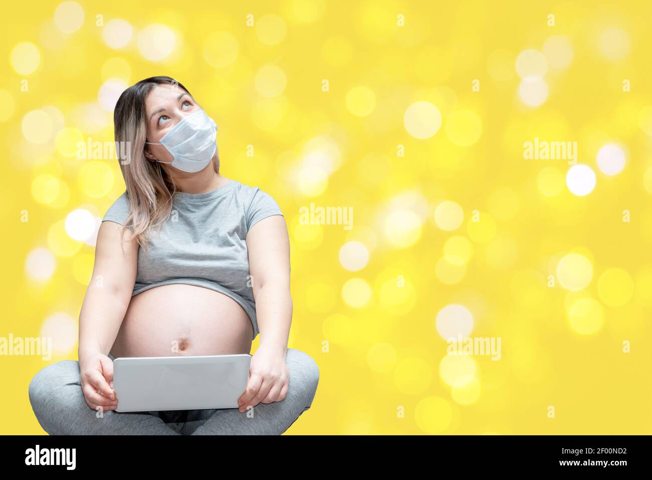 Jeune femme en espérance, portant un masque de protection et regardant en ce qui concerne l'accouchement tout en utilisant le comprimé sur jaune arrière-plan avec bokeh Banque D'Images